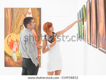 young attractive woman standing in an art gallery next to a man, pointing at some artwork displayed on wall