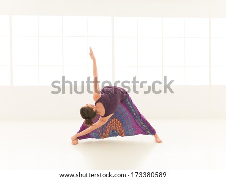 young, blonde woman demonstrating stretching yoga postrure, with face obscured, dressed colorful, iluminated window backgrond