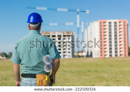 back view of male civil engineer holding plans, looking at building project in progress