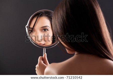 a beauty studio picture of a young woman looking in a mirror, while her back is turned to the camera. the mirror held over her left shoulder reflects her smiling face.