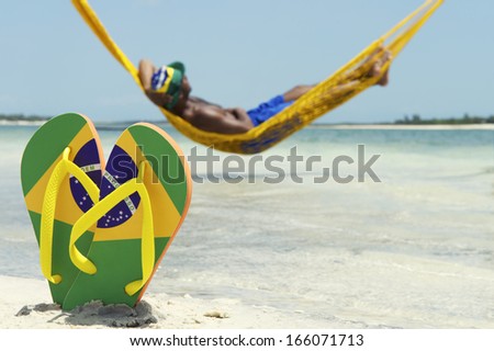 Man relaxes in hammock over the sea leaving his Brazil flag flip flops in the sand