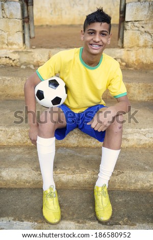 Smiling portrait of young Brazilian soccer player holding football sitting outdoors in his favela neighborhood