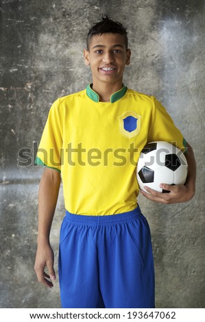 Smiling young Brazilian soccer player in team uniform of Brazil colors holding vintage football against concrete favela wall