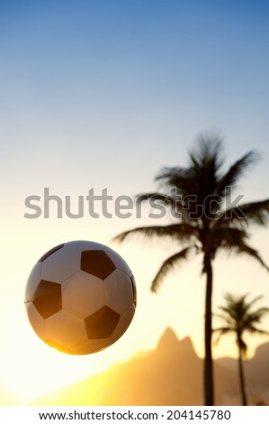 Soccer ball football flying in the golden sunset sky above Rio de Janeiro Brazil skyline with Two Brothers Mountain and palm tree silhouettes