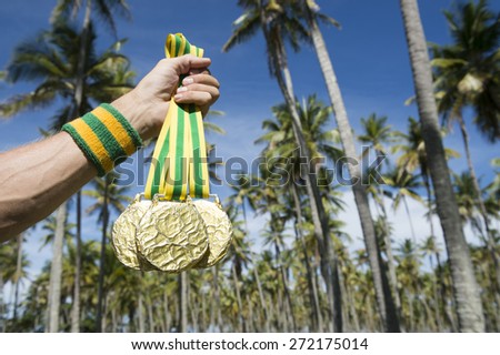 Hand of first place Olympic athlete with Brazil colors wristband holding gold medals against tropical background of palm trees