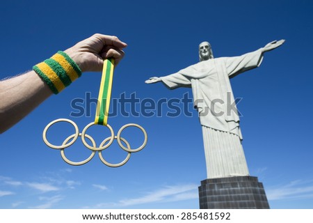 RIO DE JANEIRO, BRAZIL - CIRCA MARCH, 2015: Illustrative editorial of hand holds Olympic rings gold medal hanging from ribbon in clear blue sky next to the statue of Christ the Redeemer.