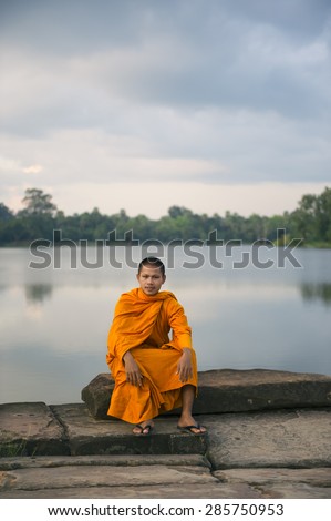 SIEM REAP, CAMBODIA - OCTOBER 30, 2014: Novice Buddhist monk in orange robe sits on the edge of the moat walkway entrance to Angkor Wat.