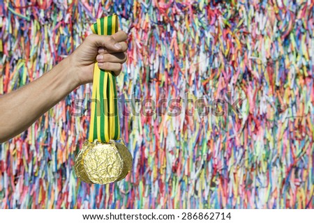 Hand holding gold medals in front of a colorful wall of good luck wish ribbons at the Bonfim church