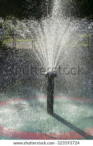Fountain spraying water backlit by dappled sun in a summer playground