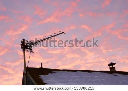 Television antenna over the snowy roof of the old house against reddish cloudy sky in evening time