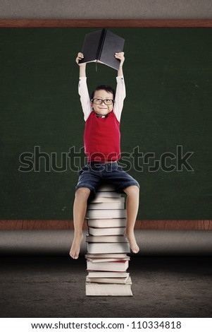 Portrait of asian schoolboy holding a book and sitting on a stack of books in front of blackboard