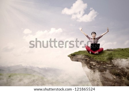 A young girl is sitting on a high mountain with her laptop and arms raised