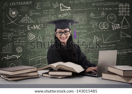 Asian female graduate is typing on laptop with books at classroom
