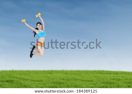 Attractive fitness woman jumping and holding dumbbells on the meadow