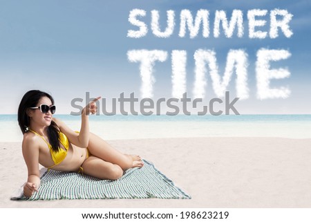 Portrait of smiling beautiful woman wearing bikini lying on mat and pointing at a text of summer time