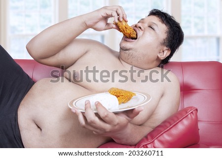 Fat man eats fast food while sitting on sofa at home