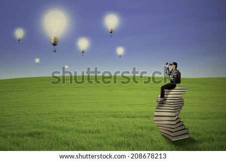Female student sitting on the stack of books while looking for future by using binoculars