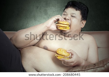 Fat man eating two hamburgers while watching tv at home