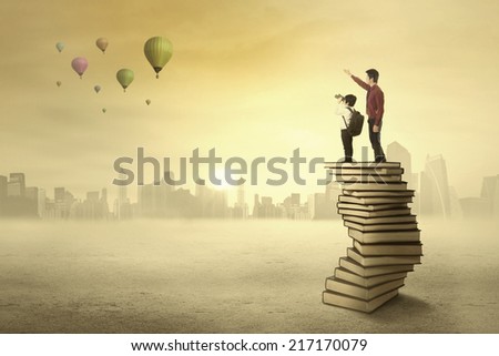 Teacher and his student standing on a pile of books looking at air balloon on the sky