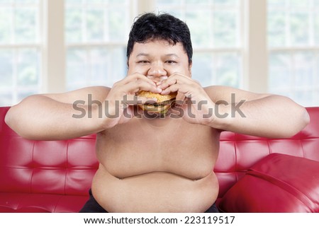 Overweight man sitting on sofa while eating hamburger at home