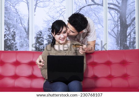 Portrait of man holding a credit card and kissing his wife on the sofa while using laptop at home