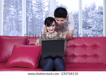 Portrait of man standing on the back of sofa and pointing on the laptop, while his wife using the laptop