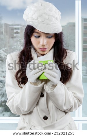 Young asian girl wearing winter jacket and standing by the window while enjoy hot drink
