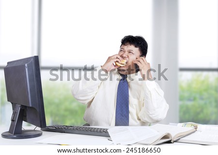 Overweight person in business suit, working in the office while eating burger and speaking on the phone