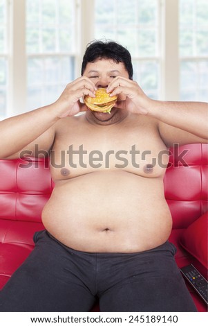 Portrait of overweight person sitting on sofa while eating burger