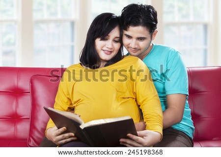 Portrait of happy pregnant couple reading a book together on the sofa at home