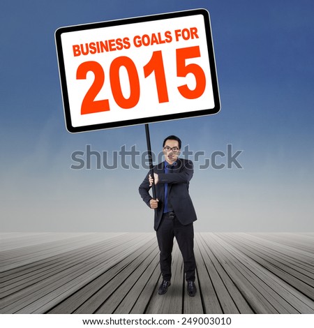 Young asian businessman standing on wooden floor while holding a board with a text of business goals for 2015