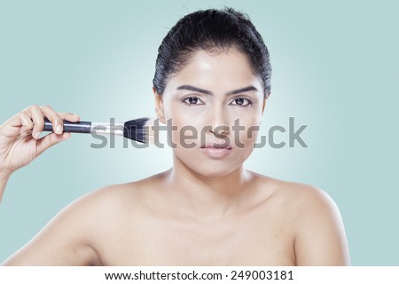 Attractive asian woman using makeup brush on her face, shot in the studio against blue background