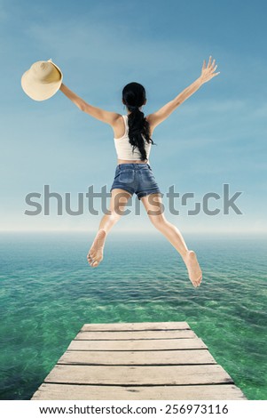 Back view of young woman holding a hat and jump on the pier to enjoy her holiday