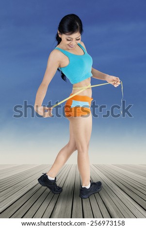 Full length of fitness woman standing on the wooden floor while measuring her waist with measuring tape