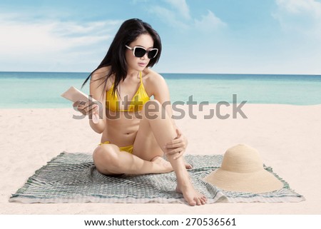 Gorgeous young asian woman sitting at beach while wearing sexy bikini and using sun cream