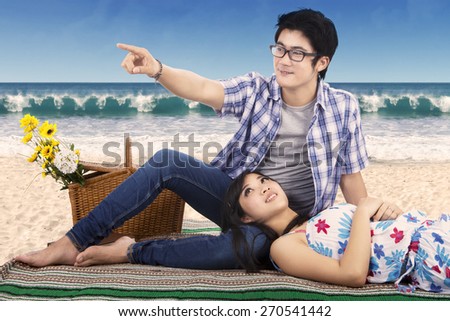 Attractive young couple enjoy holiday while sitting on mat at beach and looking at something