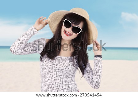 Portrait of excited young woman with long hair, smiling at the camera while wearing sunglasses and hat on the beach