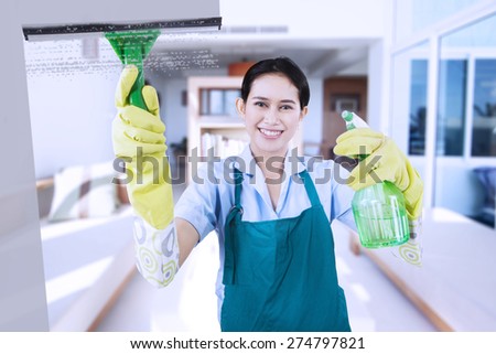Portrait of friendly maid wearing uniform and apron, cleaning a mirror with a spray while smiling