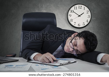 Caucasian businessman sleeping on the tablet with a clock on the wall, shot at workplace