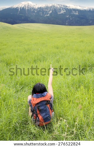 Rear view of female hiker sitting on the green grass while carrying backpack and pointing on the mountain view