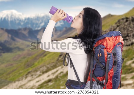 Attractive young backpacker drinking fresh water while carrying backpack for hiking on the mountainside