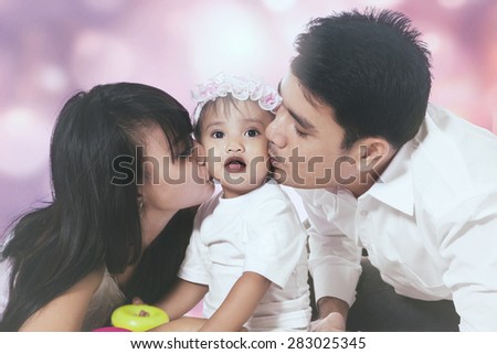 Portrait of two parents kissing their baby together, shot against a light glitter background