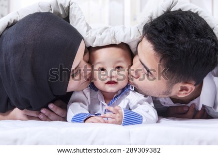 Portrait of two young muslim parents lying on the bed while kissing their baby under the blanket