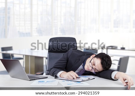 Male caucasian worker wearing glasses in office and sleeping on desk with laptop and paper works