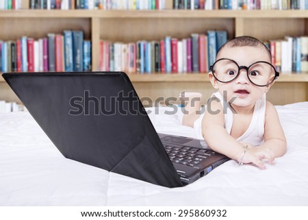 Cute baby lying on bed while wearing glasses with laptop and a bookshelf background