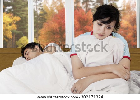 Portrait of unhappy young woman sitting on bed near her husband with autumn background on the window