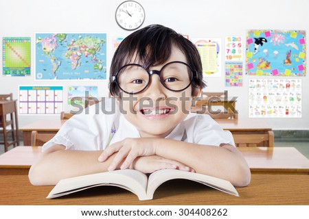 Excited female elementary school student wearing glasses in the class and smiling at the camera while reading a book on the table