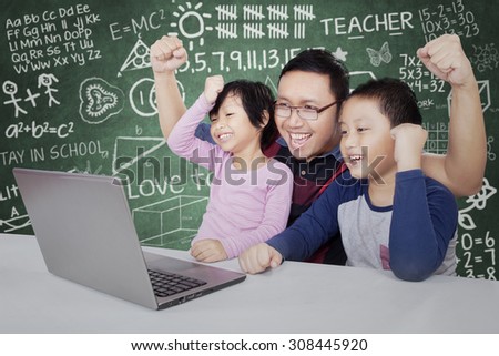 Two attractive students and their teacher celebrate success by raising hands together while looking at the laptop computer in the classroom