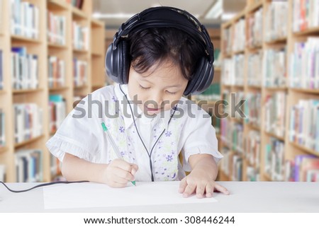 Female primary school student studying in the library while wearing headphones and write on the paper