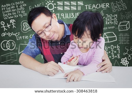 Cute female elementary school student try to write on the book with male teacher in the classroom
